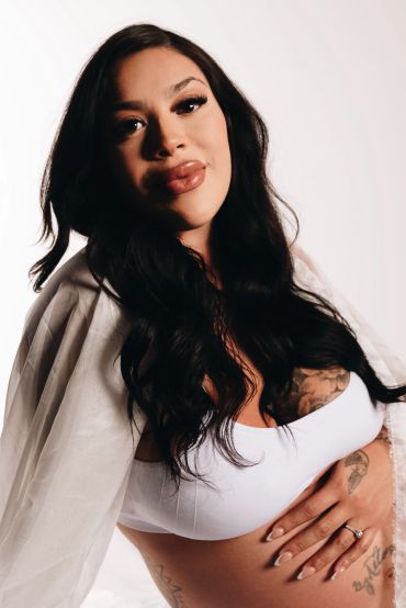 A confident woman with long black hair and tattoos poses in a white top and sheer cover-up.
