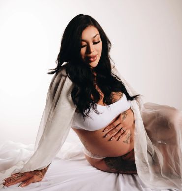 A pregnant woman with tattoos poses gracefully in white lingerie and a sheer robe.