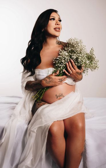 Pregnant woman with tattoos holds a bouquet of white flowers, wrapped in a sheer white fabric.