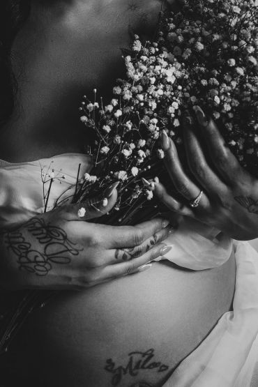 Black and white photo of a tattooed person holding flowers over their pregnant belly.