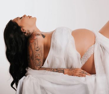 Pregnant woman with tattoos lying back, draped in white fabric.