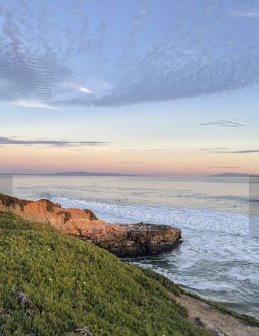 Coastal cliff with surfers in the ocean under a pastel sunset sky.