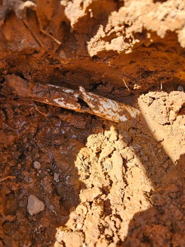Broken white pipe buried in wet, muddy soil with sunlight casting shadows.