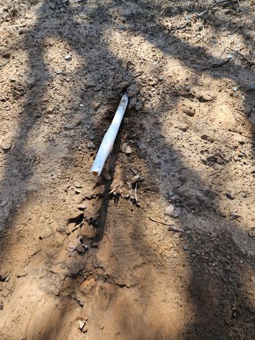 A white pipe partially buried in dirt with shadows overhead.