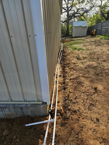 Two white PVC pipes along a building's side leading to a small shed in a yard.