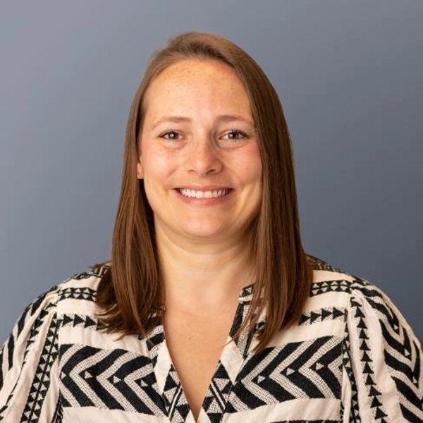 Smiling woman with straight brown hair in a patterned black and white top against a gray background.