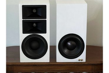 Two white speakers with black drivers on a wooden surface against a beige wall.