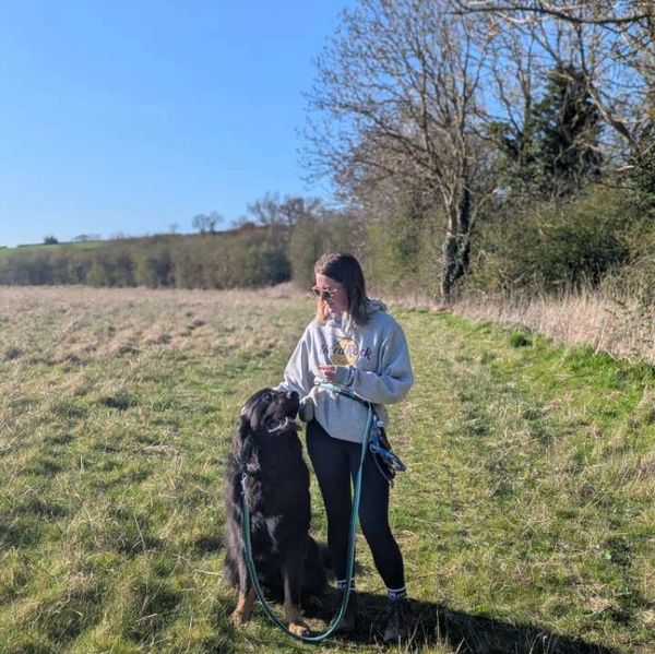 Woman in hoodie walking a large black dog on a sunny grassy path.