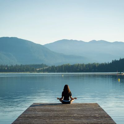 A person meditating on a dock by a calm lake surrounded by mountains.
