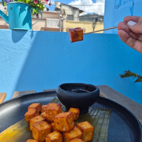 Skewered tofu cube held above a black plate with more tofu and dipping sauce.