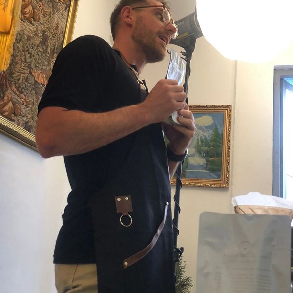 Man in apron holding a package, seemingly explaining something indoors.