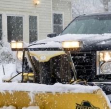 Black pickup truck with yellow snow plow clearing snow in front of a house.
