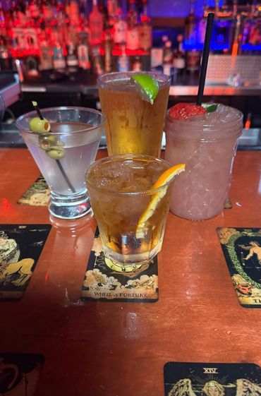 Four colorful cocktails with fruit garnishes on tarot card coasters at the bar.