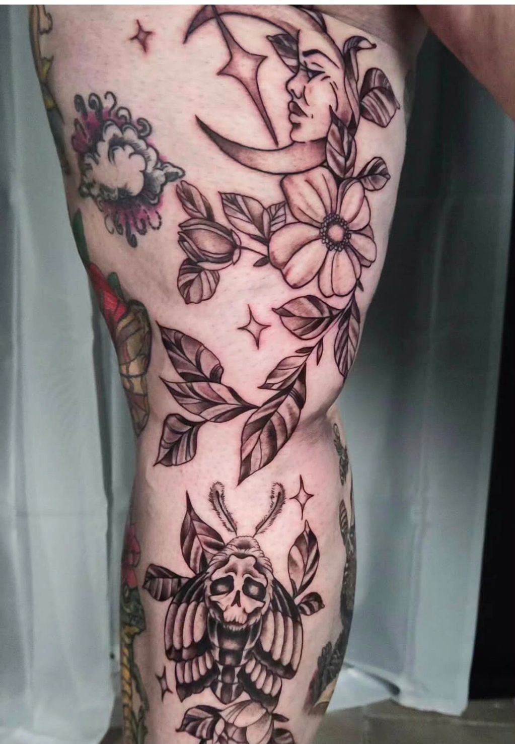 Black and gray tattoo of moon, flowers, and a skull moth on leg.