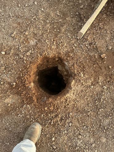 A freshly dug hole in rocky soil with a boot and wooden stick nearby.