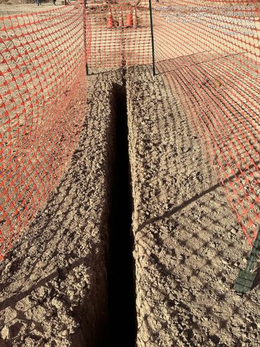 A deep, narrow trench is fenced off with orange safety netting on a dry, dusty construction site.