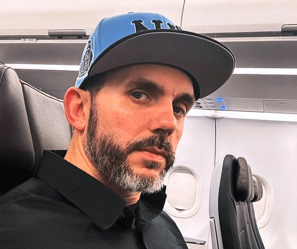 Man with a beard wearing a blue cap sitting on an airplane.