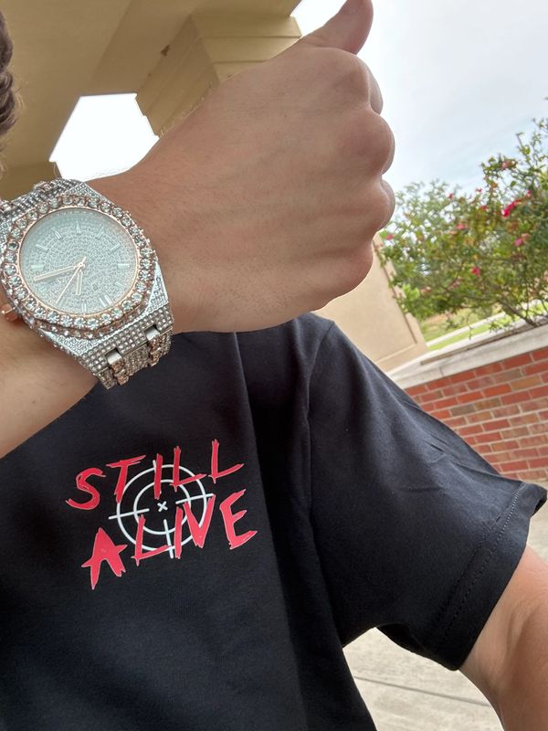 Person wearing a sparkling watch and a "Still Alive" black t-shirt giving a thumbs up.