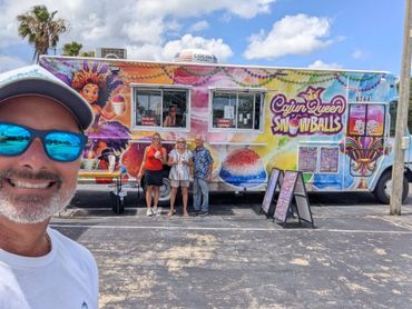 Man taking selfie in front of colorful snowball truck with people standing nearby.