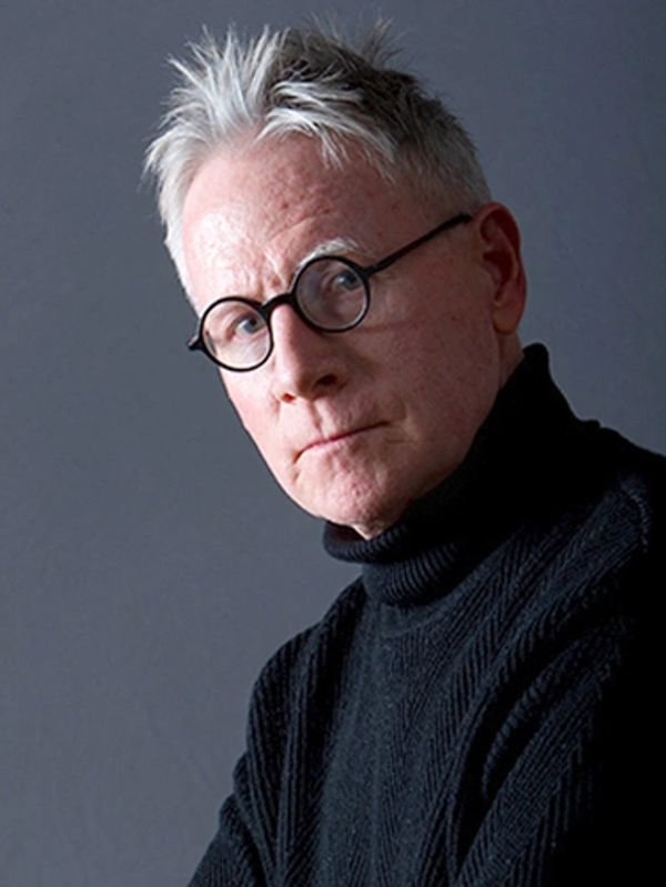 Serious elderly man with round glasses and white hair in black turtleneck.