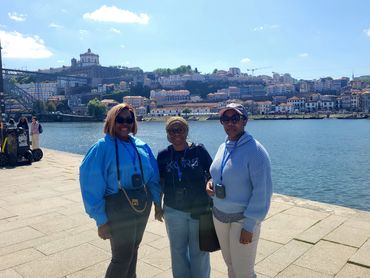 Three women posing by a river with a scenic city and bridge in the background on a sunny day.