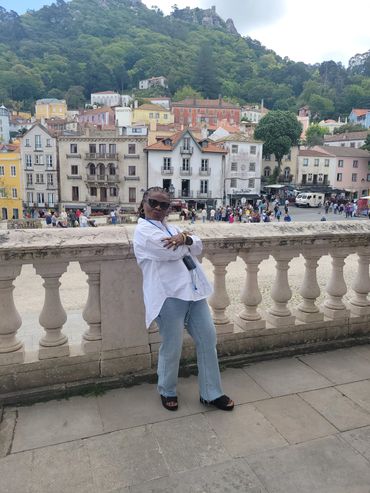 Woman posing confidently on a stone balcony with colorful buildings and green hills in the background.
