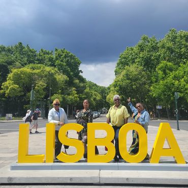 Four women posing behind large yellow LISBOA letters under a cloudy sky.