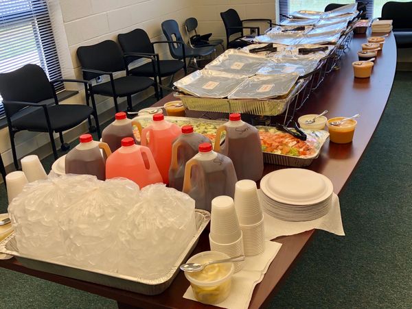 Buffet setup with drinks, plates, and covered food trays on a conference table.