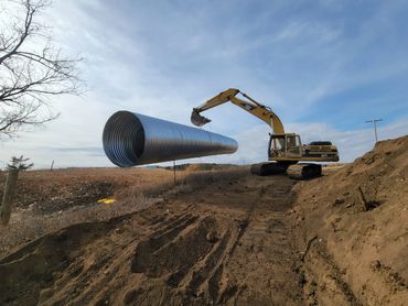 Excavator lifting a large metal pipe on a construction site.