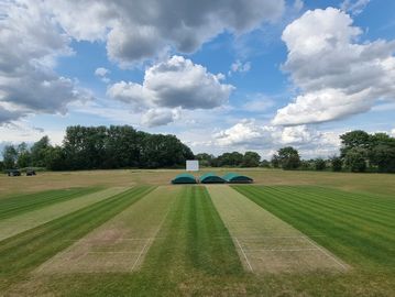 A cricket pitch under a partly cloudy sky with green grass and trees in the background.