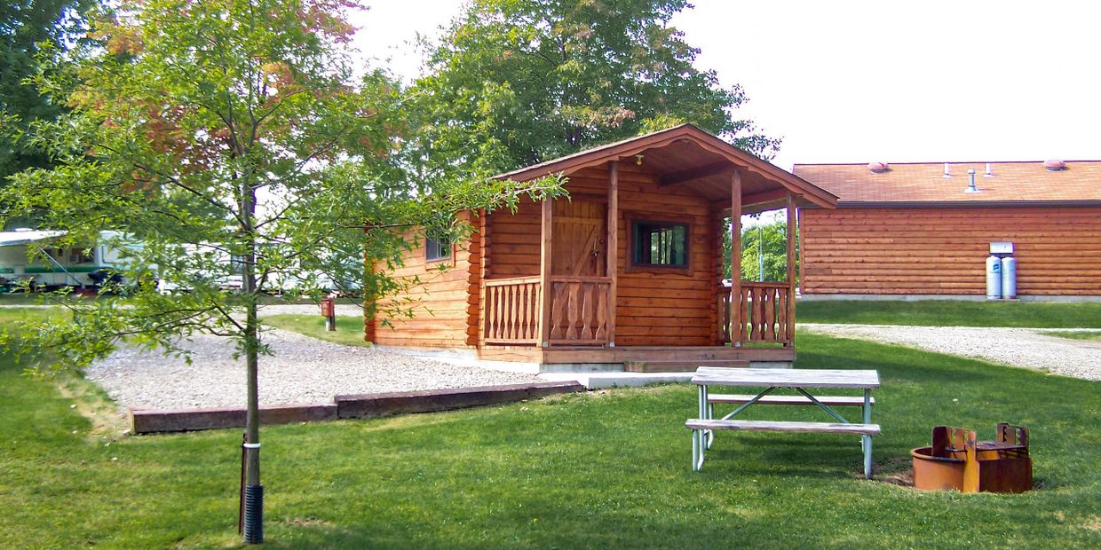 Log cabin with gravel parking beside and picnic table and fire ring in front.