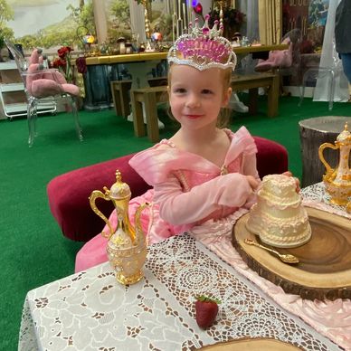 A young girl in a pink princess dress and birthday tiara enjoys a tea party with ornate decorations.