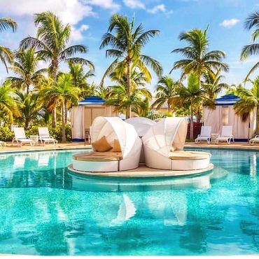 Luxury resort pool with round white lounge chairs, surrounded by palm trees and cabanas under blue