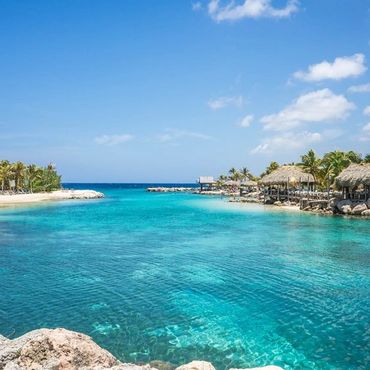 Tropical lagoon with clear turquoise water, palm trees, and thatched huts under blue sky.
