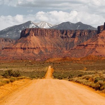 Dirt road leading through desert landscape with red rock mesas and snow-capped mountains.