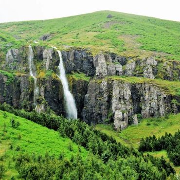 Waterfall cascading down rocky cliffs surrounded by lush green hills and trees.