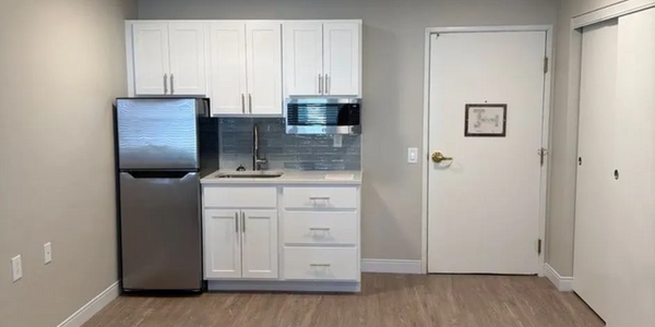 Compact kitchenette with white cabinets, stainless steel fridge, microwave, and wood flooring.