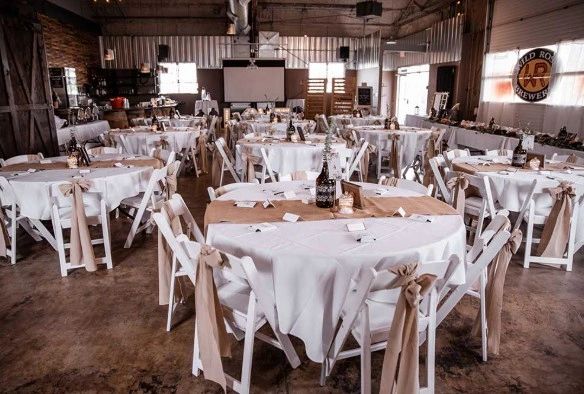 Rustic event space with round tables and white chairs decorated with beige bows.
