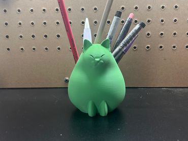 Green cat-shaped pen holder with various pens and pencils on a black surface.