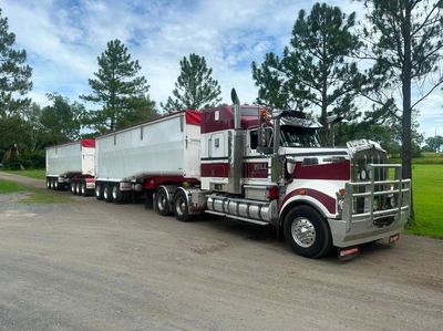 A large red and white semi-truck with two trailers parked on a road beside trees.