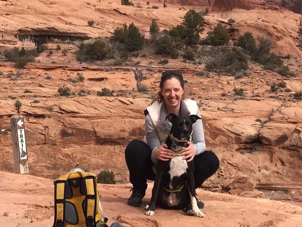 A woman crouches with her dog on red rock terrain near a large natural sandstone arch.