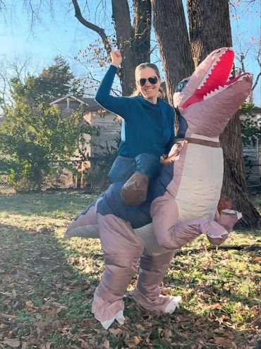 Woman in a dinosaur costume, smiling and posing in a sunny backyard with trees.