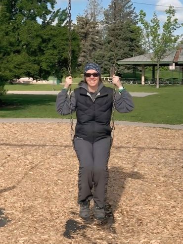 Woman sitting on a swing in a park, wearing sunglasses, a beanie, and a vest.
