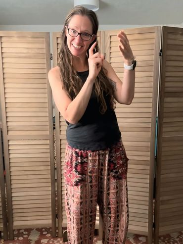 A smiling woman in a black tank top and patterned pants gestures playfully in a cozy room.