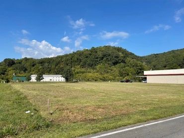 Open grassy field with buildings and wooded hills under a blue sky.