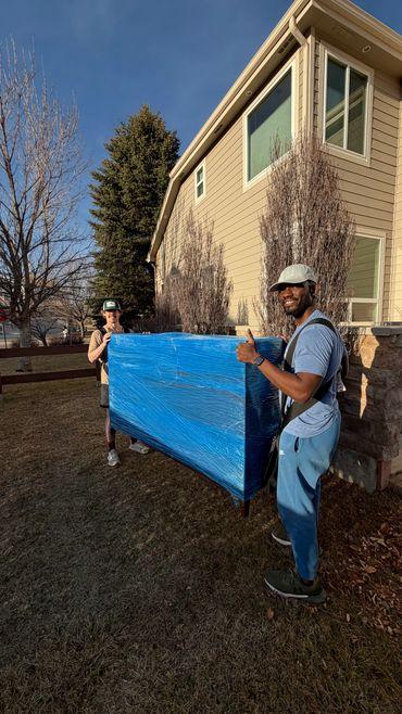 Two men carrying a large blue-wrapped item outside a house, one giving a thumbs-up.
