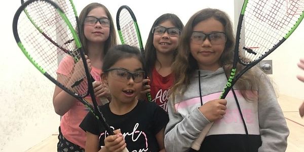 four girls posing with squash racquets in a squash court wearing safety goggles 