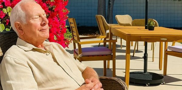 Elderly man relaxing in the sun beside a bright flower bush.
