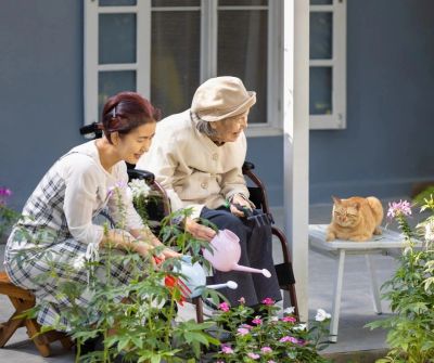 Caregiver providing companion and supervision to elderly woman while watering flowers outside.