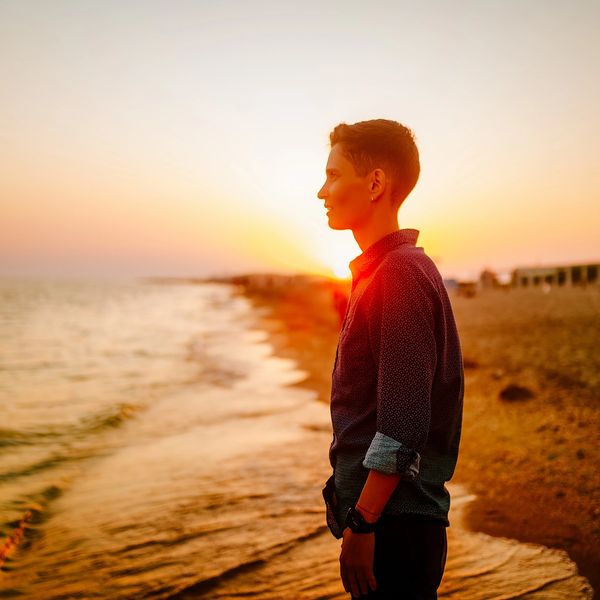 A person stands on a beach looking out at the sea with the sun setting behind them
